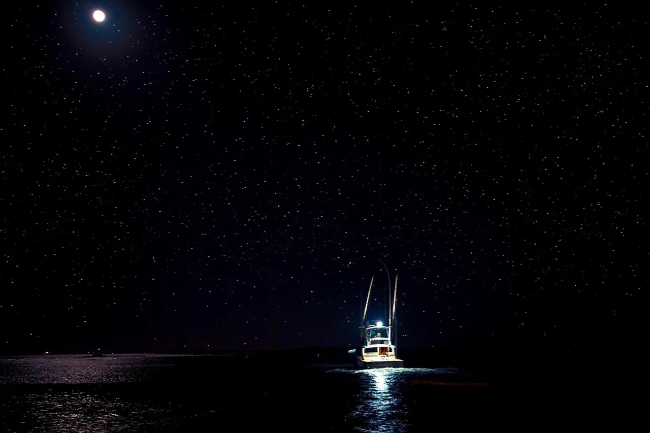 A sportfishing boat at anchor at night under a full moon, its deck lit by floodlights